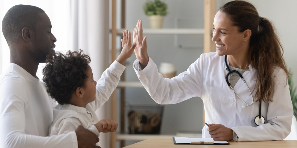 Photo of a white woman doctor giving a positive â€œhigh fiveâ€ hand gesture to a young patient as he sits in his fatherâ€™s lap.